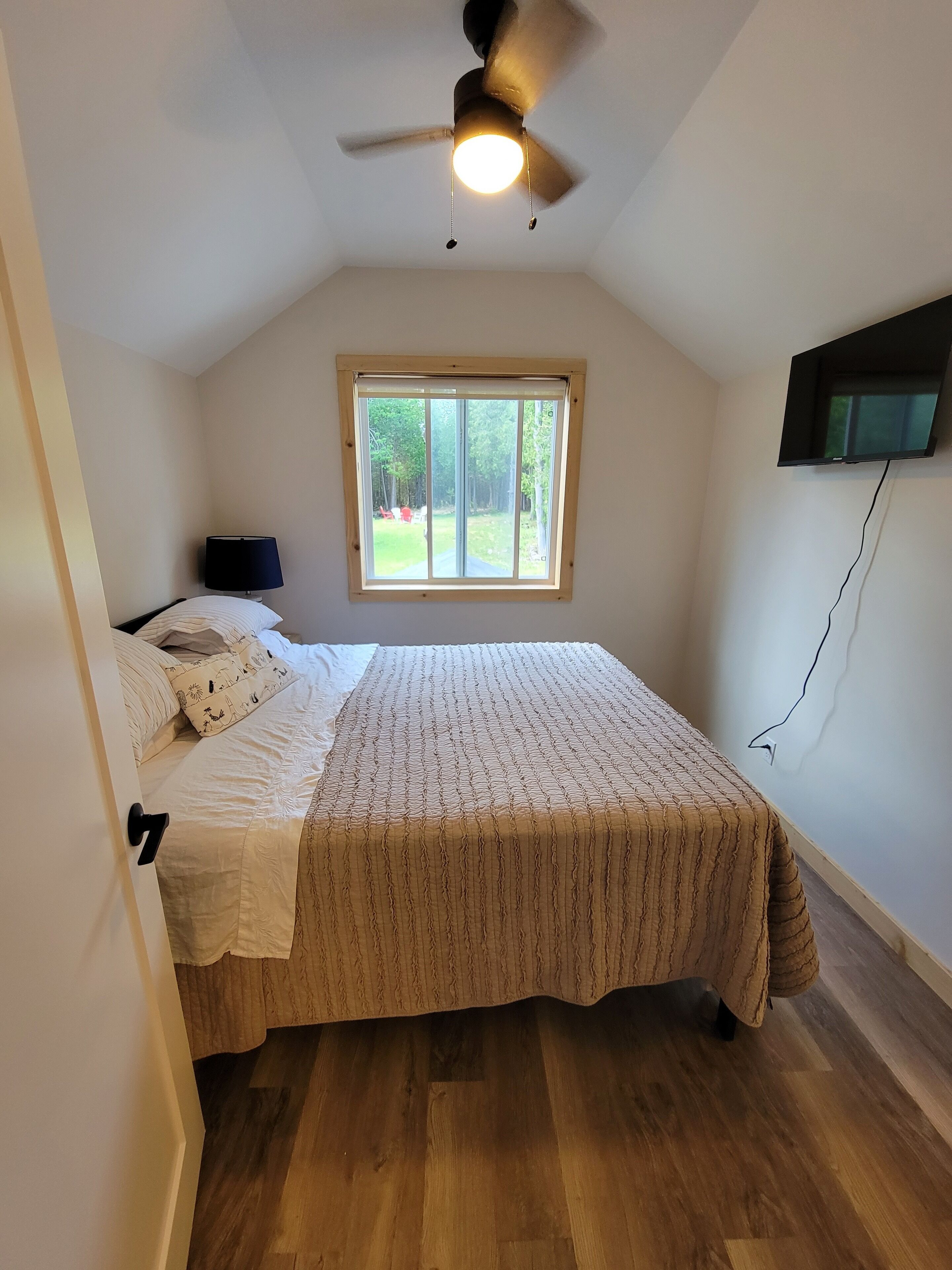 A bedroom with a bed, a ceiling fan, a window, and a TV mounted on the wall.