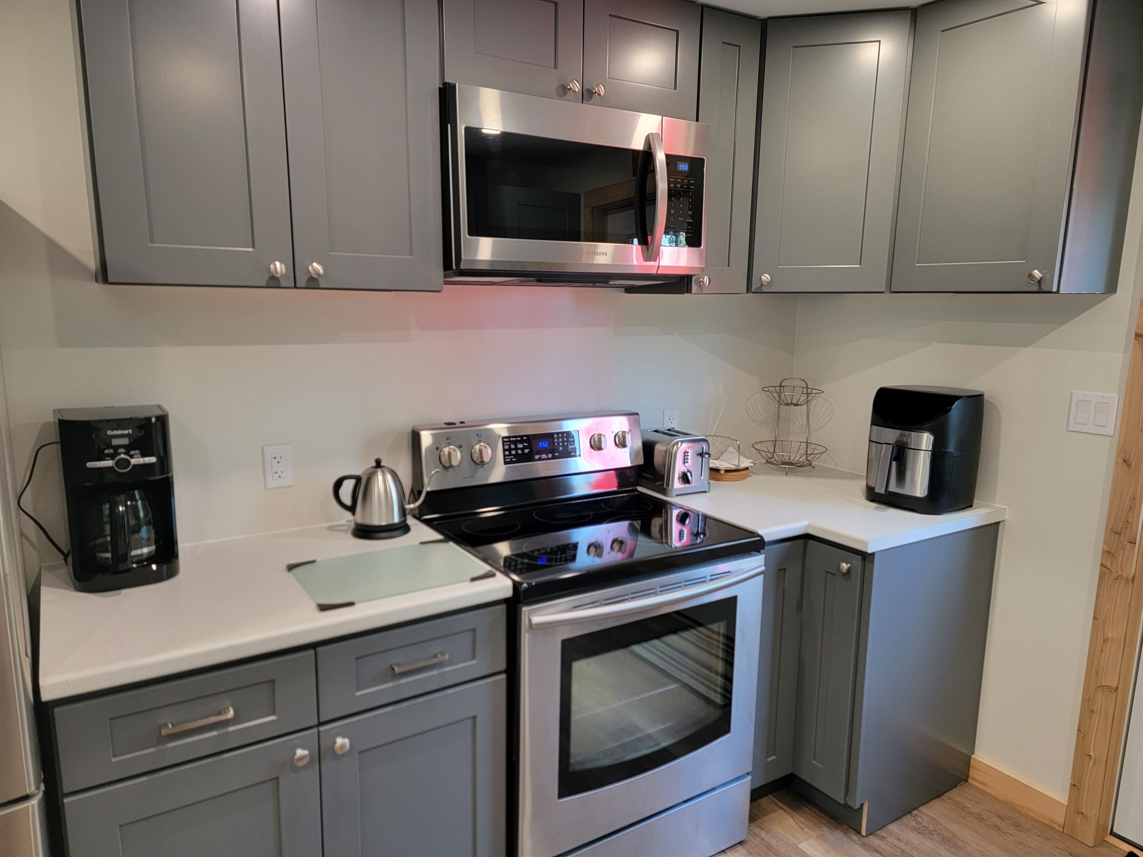 A modern kitchen with grey cabinets, a stainless steel microwave, a coffee maker, a kettle, a toaster, and an oven.