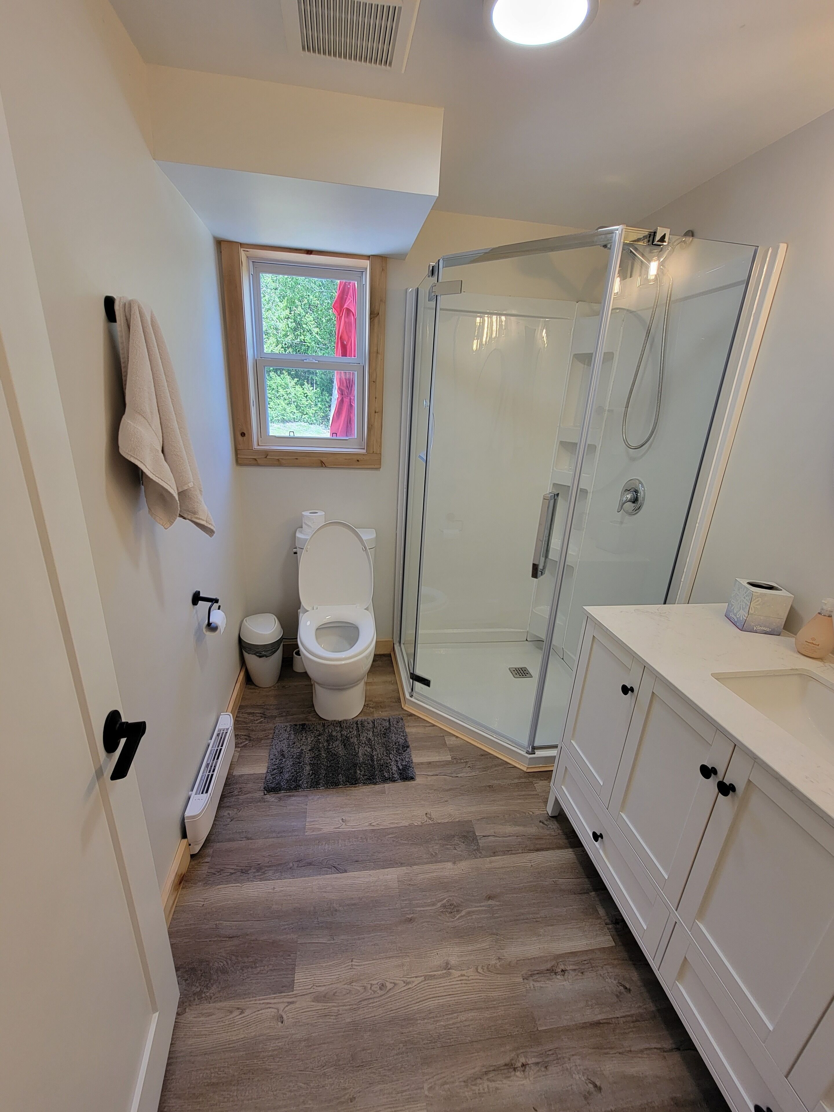 A bathroom with a glass shower enclosure, a toilet, a sink, and a window with a view of greenery.
