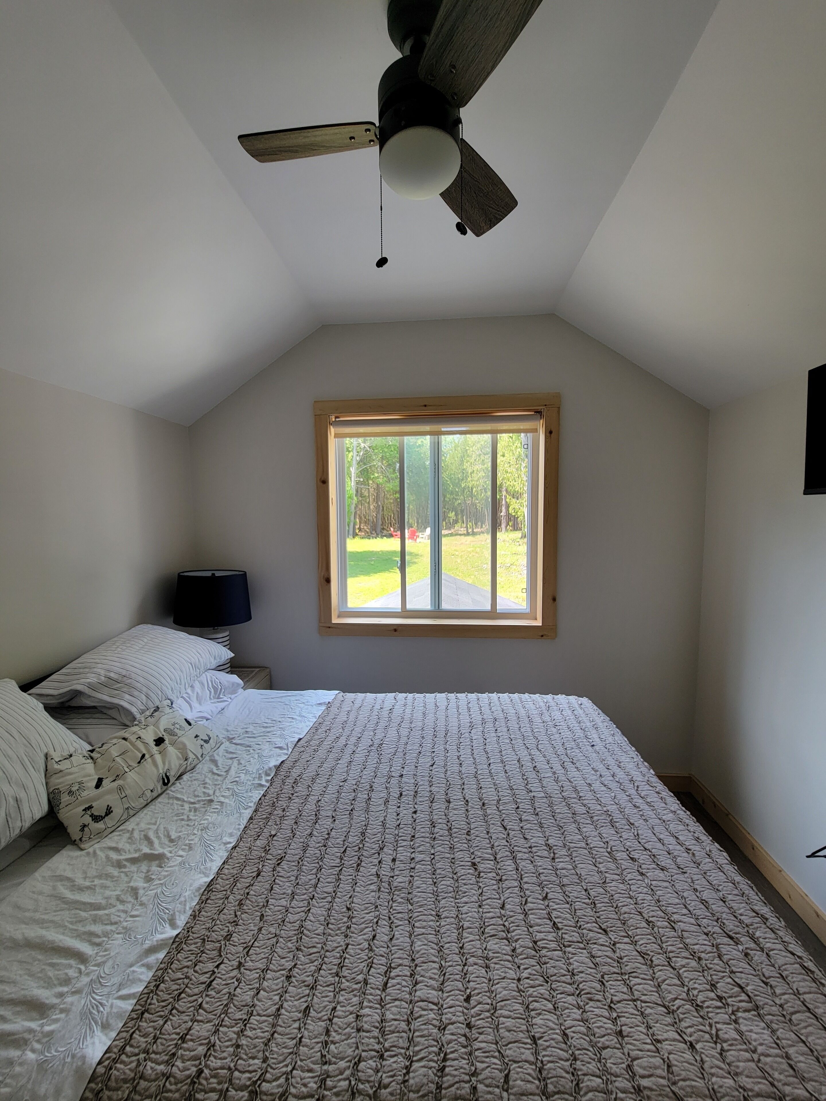 A bedroom with a large bed, a ceiling fan, a window with a view of the outdoors, and a nightstand with a lamp.