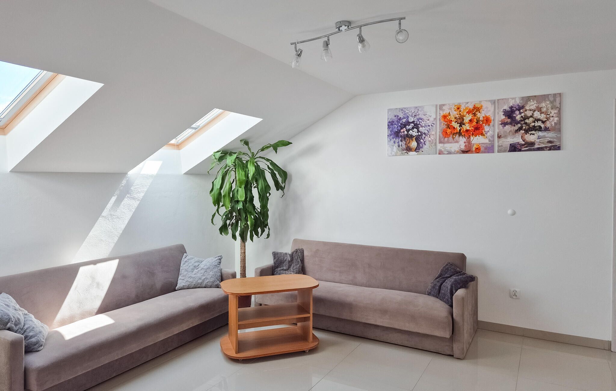 Un salon moderne avec un plafond en pente, une grande fenêtre, un canapé gris, une table basse en bois, une plante en pot et une série de tableaux floraux accrochés au mur.