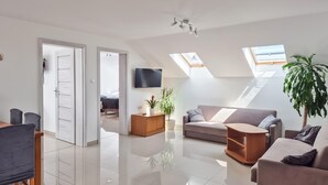 A modern living room with a sofa, coffee table, and a TV. There are two potted plants and a wooden cabinet. The room has a high ceiling with skylights and a tiled floor. - Awesome apartment in Dobrzyca with WiFi (Dobrzyca)