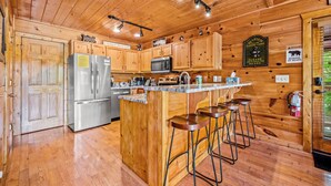 A wooden cabin kitchen with a stainless steel refrigerator, a microwave, a sink, and bar stools. - Rustic Pigeon Forge Cabin - Mountain View/King Beds/Hot Tub/Minutes to Dollywood (Sevierville)