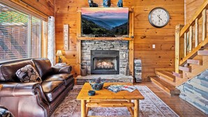 A cozy cabin interior with a leather couch, a fireplace, a stone fireplace surround, a wooden table with magazines, and a clock on the wall. - Rustic Pigeon Forge Cabin - Mountain View/King Beds/Hot Tub/Minutes to Dollywood (Sevierville)