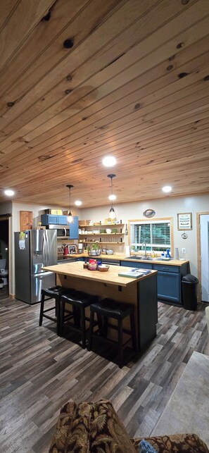 Une cuisine avec un plafond en bois, des appareils en acier inoxydable, un îlot central avec des tabourets et une fenêtre avec des stores.