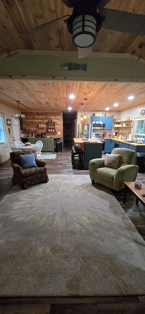 A spacious living area with a wooden ceiling, a dining table with chairs, and a kitchen in the background.