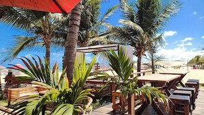 Eine Terrasse direkt am Strand mit Holztischen und -stühlen, umgeben von Palmen und blauem Himmel.