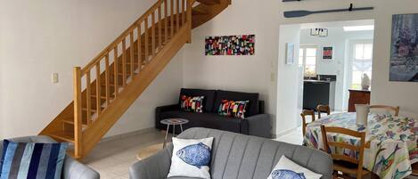 A living room with a grey sofa, a wooden staircase, a dining area with a table and chairs, and a kitchen visible through an open doorway.