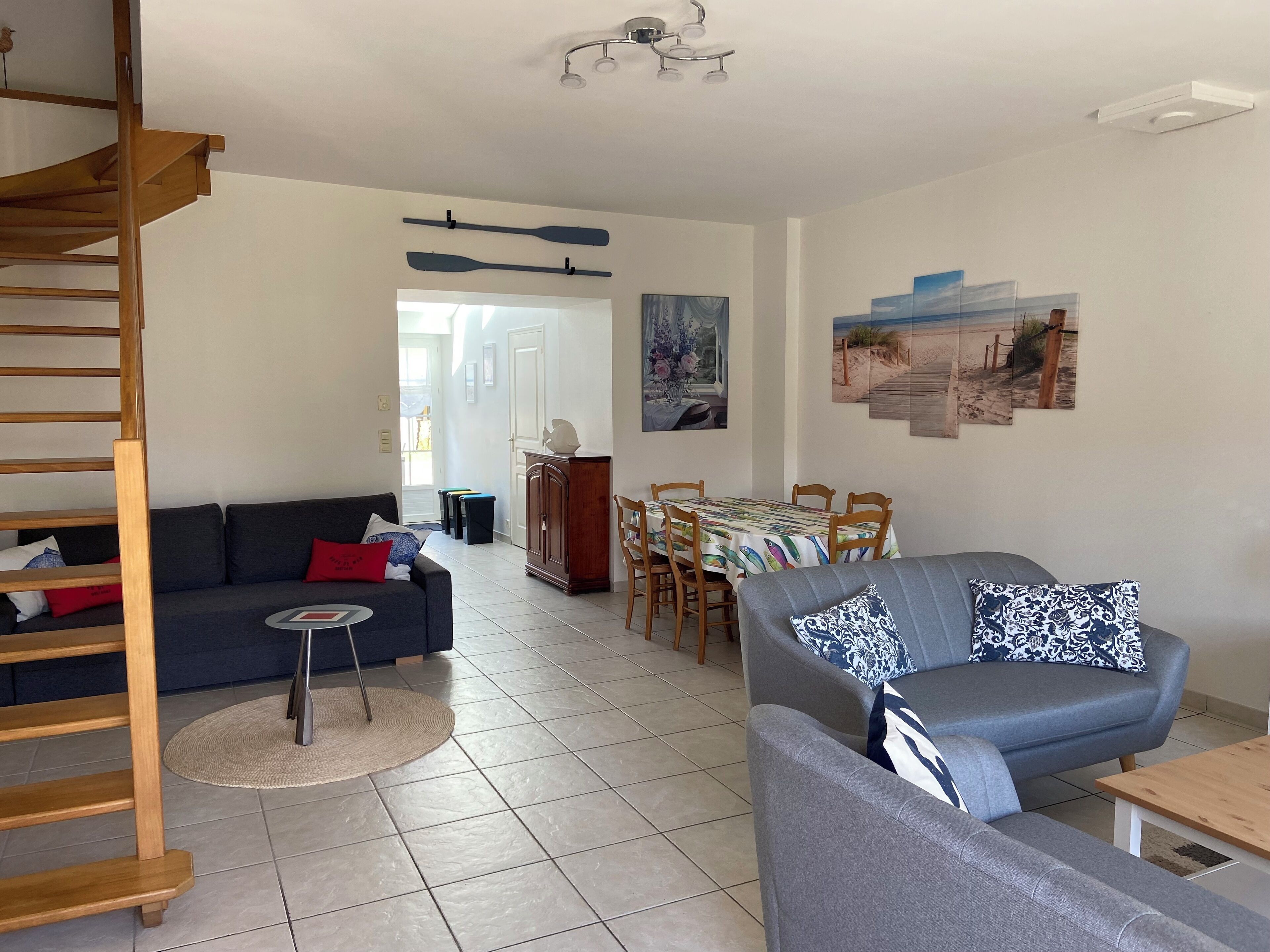 A living room with a grey sofa, a wooden staircase, a dining area with a table and chairs, and a painting of a beach scene on the wall.