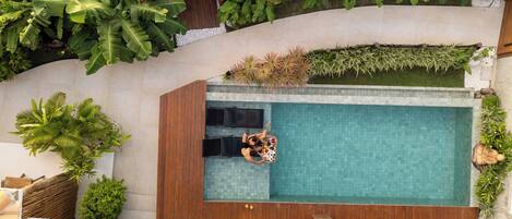 A rooftop pool with two lounge chairs, surrounded by lush greenery and a wooden deck.