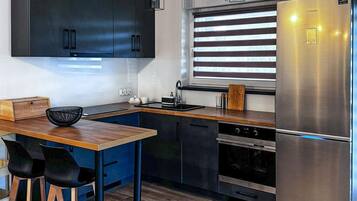 Une cuisine moderne avec des meubles de couleur sombre, un réfrigérateur en acier inoxydable, un plan de travail en bois et une fenêtre équipée de stores.
