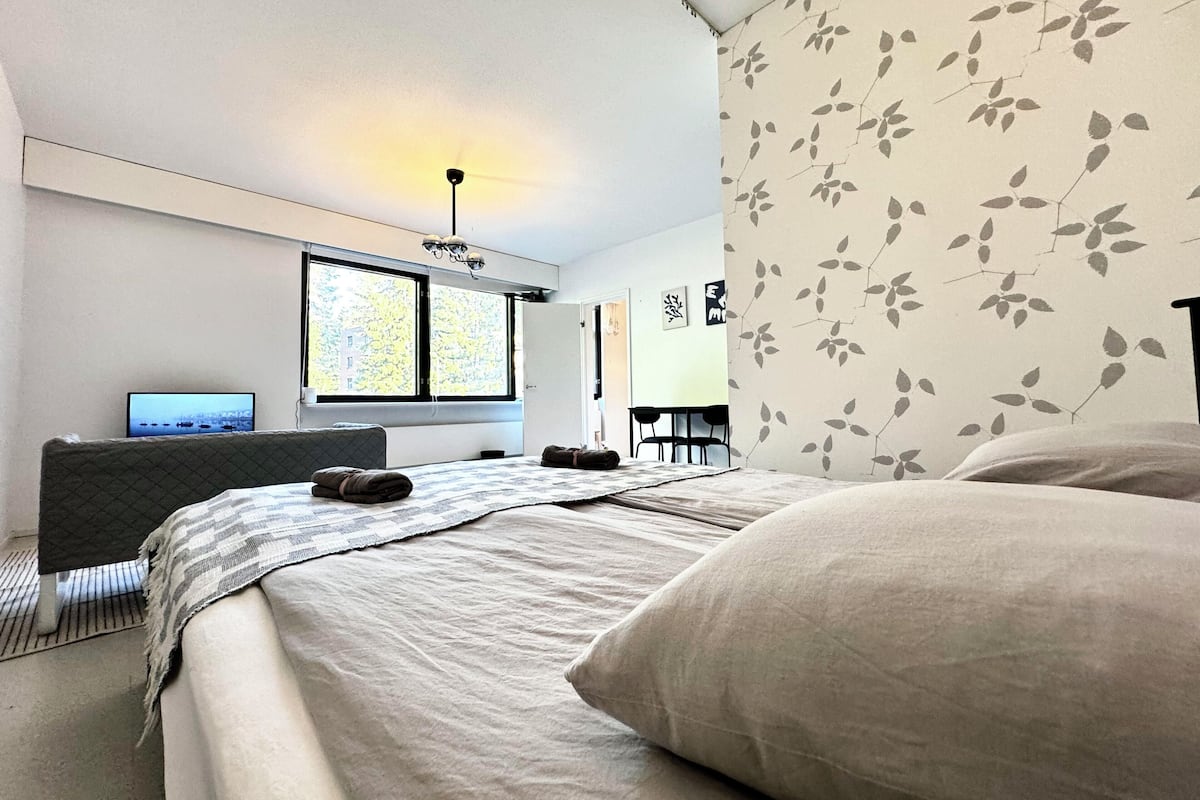 Quarto moderno com cama, escrivaninha e cadeira. Há uma janela com vista para árvores e um edifício.