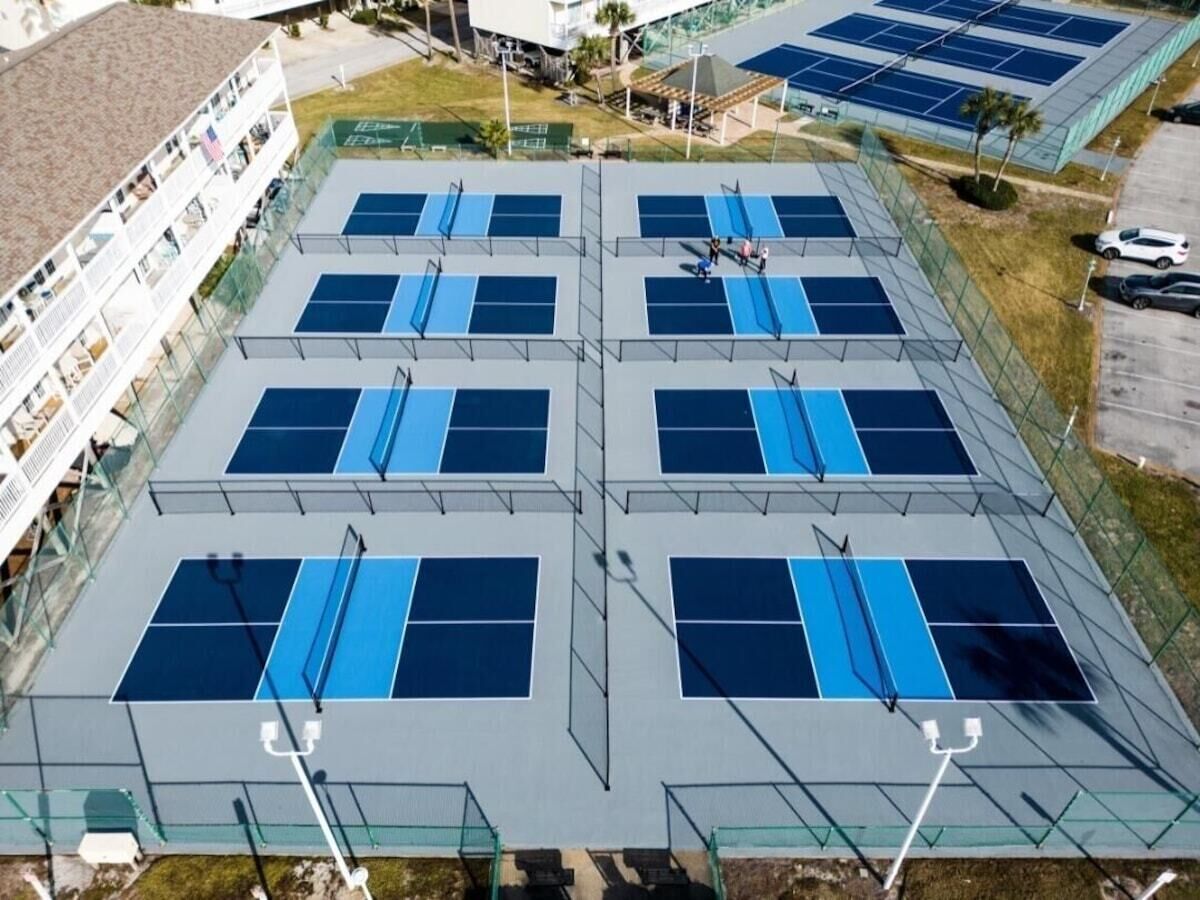 Complejo de canchas de tenis al aire libre con múltiples canchas, rodeado por una cerca y contiguo a un edificio con balcones.