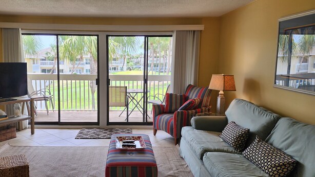 A living room with a TV, sofa, armchair, and a view of a pool and palm trees. - 2/2, Sleeps 6, 9 hole golf course on site, pool out your front door, 8 min.beach (Destin)
