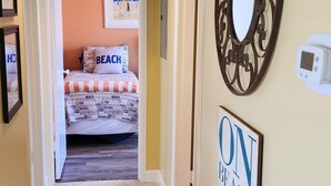 A hallway leading to a bedroom with a beach-themed bedspread, a decorative mirror, and a sign that reads "ON BEACH TIME". - 2/2, Sleeps 6, 9 hole golf course on site, pool out your front door, 8 min.beach (Destin)