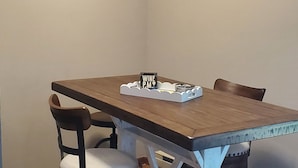 Uma área de refeições com uma mesa de madeira, duas cadeiras e um banquinho de bar. Há duas pinturas nas paredes.