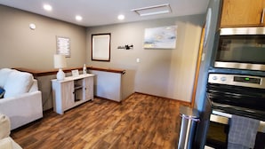 Quarto de hotel compacto com piso de madeira, um sofá branco com almofada azul, uma pequena mesa lateral branca com luminária, uma moldura com imagem na parede e uma cozinha compacta com forno de aço inoxidável.