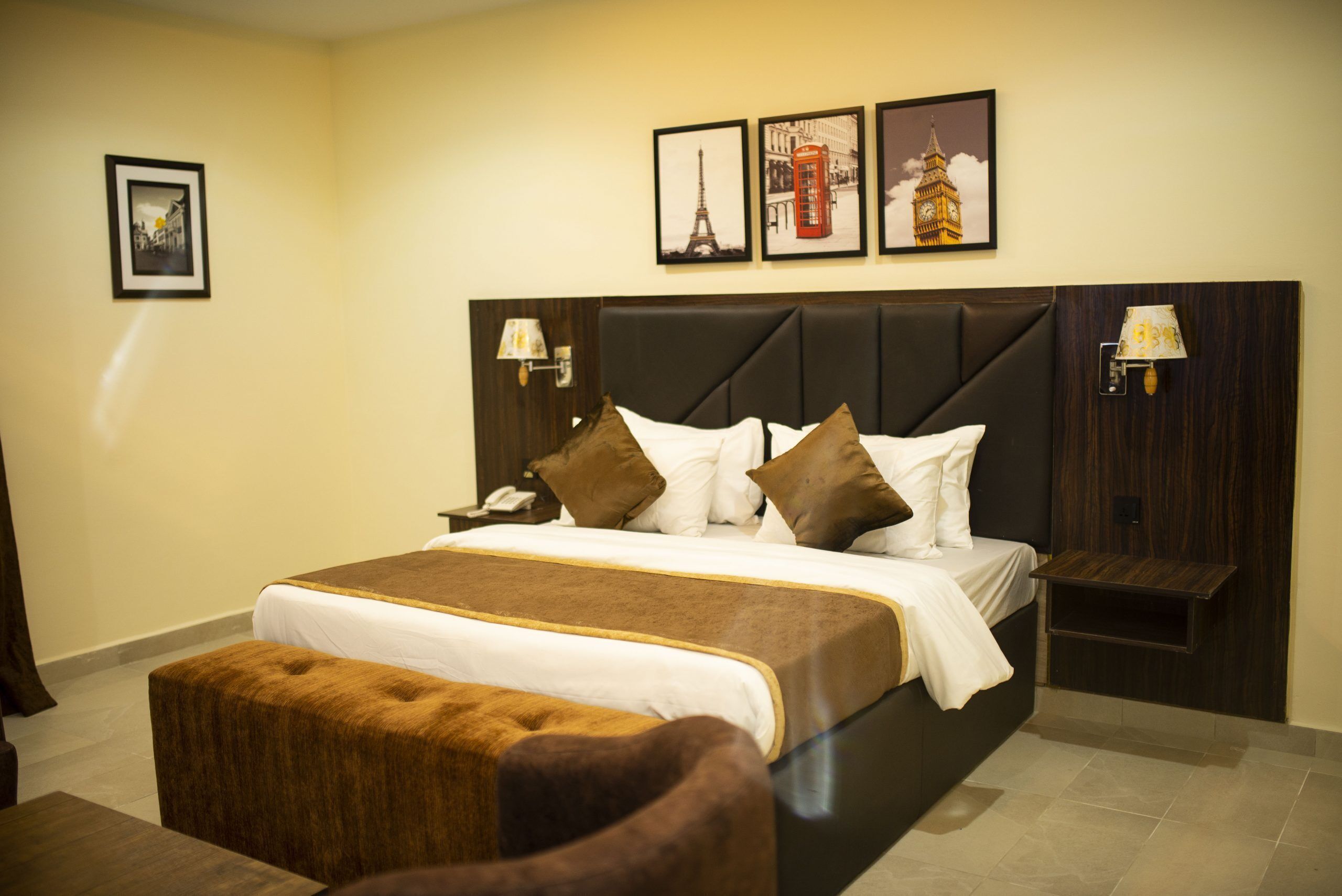 Une chambre d’hôtel avec un grand lit, une table de chevet, une lampe et une petite table. Trois tableaux encadrés sont accrochés au mur, au-dessus du lit.