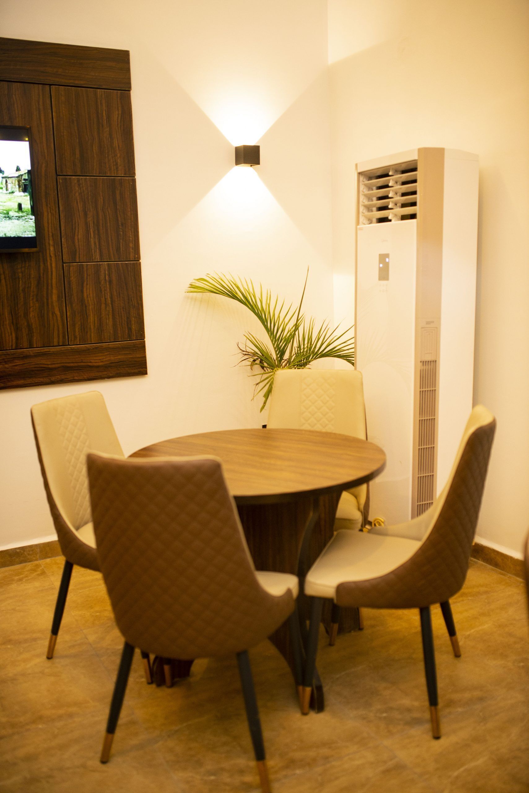A dining area with a round table, four chairs, a potted plant, a wall-mounted TV, and a built-in air conditioning unit.