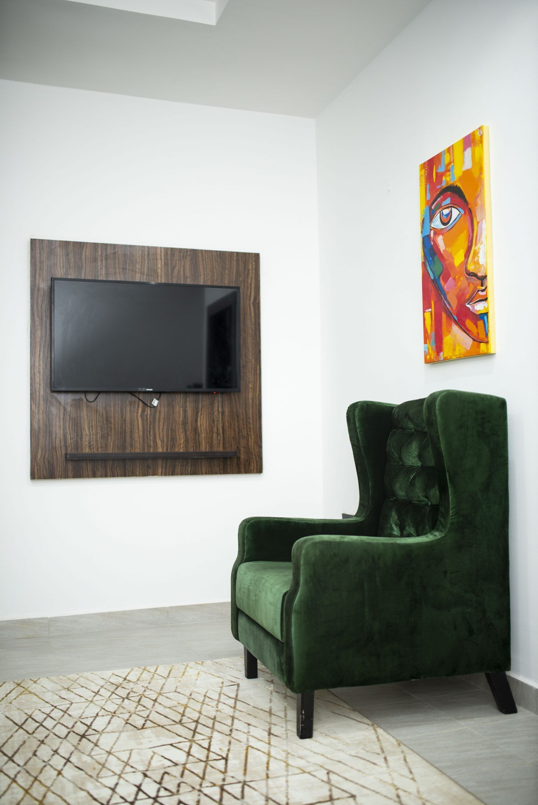 Una sala moderna con un televisor de pantalla plana montado en un panel de madera, un sillón de terciopelo verde, un cuadro abstracto de colores y una alfombra con estampado geométrico.