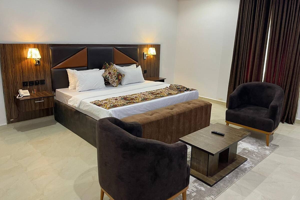A modern hotel room with a large bed, two bedside lamps, a wooden nightstand, a small table, two armchairs, and a large window with curtains.