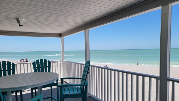 Un espacio al aire libre cubierto con una mesa y sillas, con vista a una playa donde hay gente y una persona pescando.