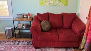 Ein rotes Sofa mit einem gemusterten Kissen, ein kleiner Tisch mit Dekoration und zwei Bilder an der Wand.