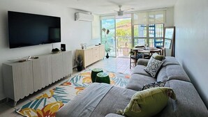 A modern living room with a grey sofa, a flat-screen TV, a ceiling fan, and a view of a balcony with a table and chairs.
