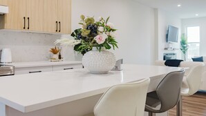 Een moderne keuken met witte kastjes, een wit marmeren aanrechtblad en een centraal eiland met een bloevas. Er zijn twee witte barkrukken en een flatscreen televisie aan de muur bevestigd.