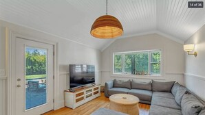 Ein gemütliches Wohnzimmer mit Fernseher, einer Couch, einem Korbtisch und einem großen Fenster mit Jalousien.