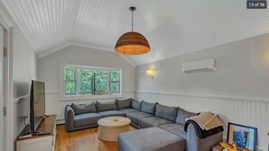 Ein modernes Wohnzimmer mit einem Ecksofa, einem runden Couchtisch, einem Flachbildfernseher und einem großen Fenster mit Blick auf Grünflächen.