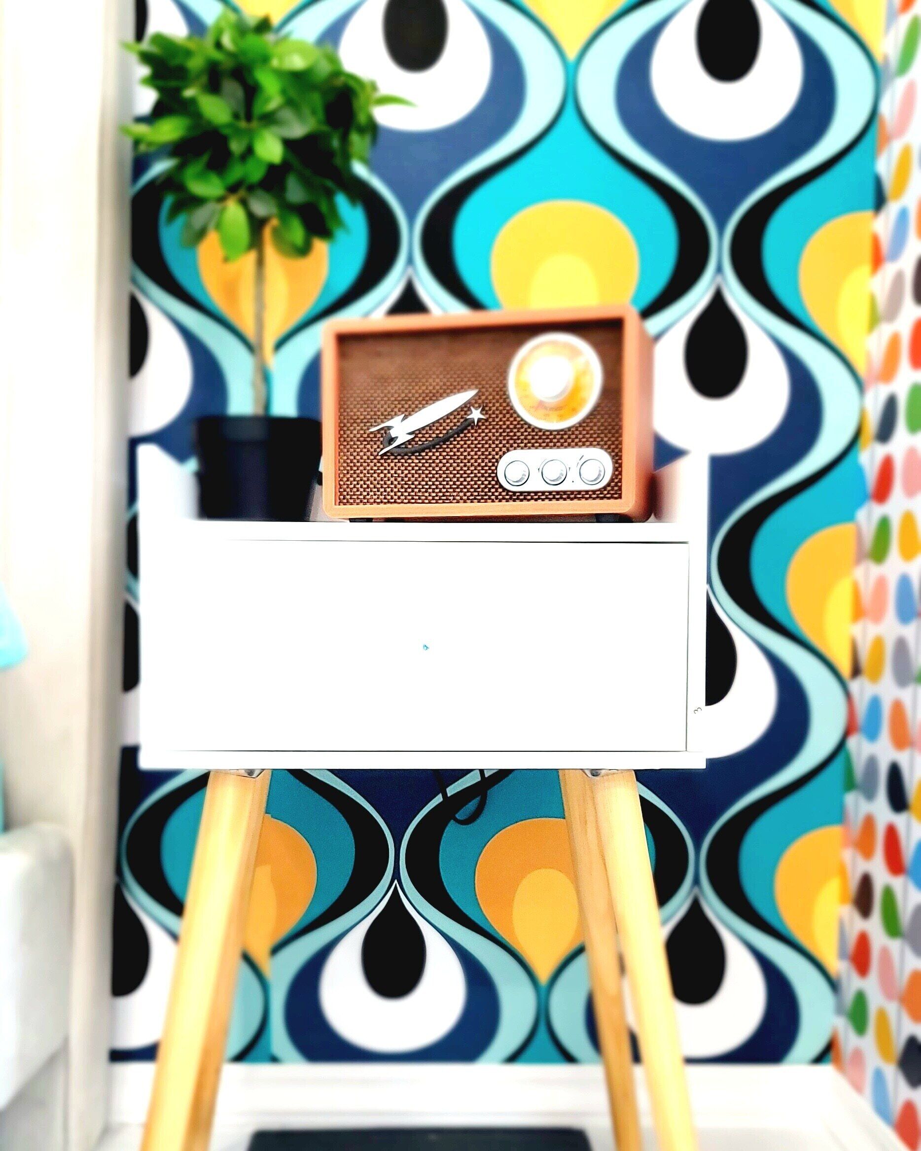 Une petite table blanche sur laquelle repose une radio ancienne, le tout placé devant un papier peint aux motifs géométriques et aux couleurs vives.
