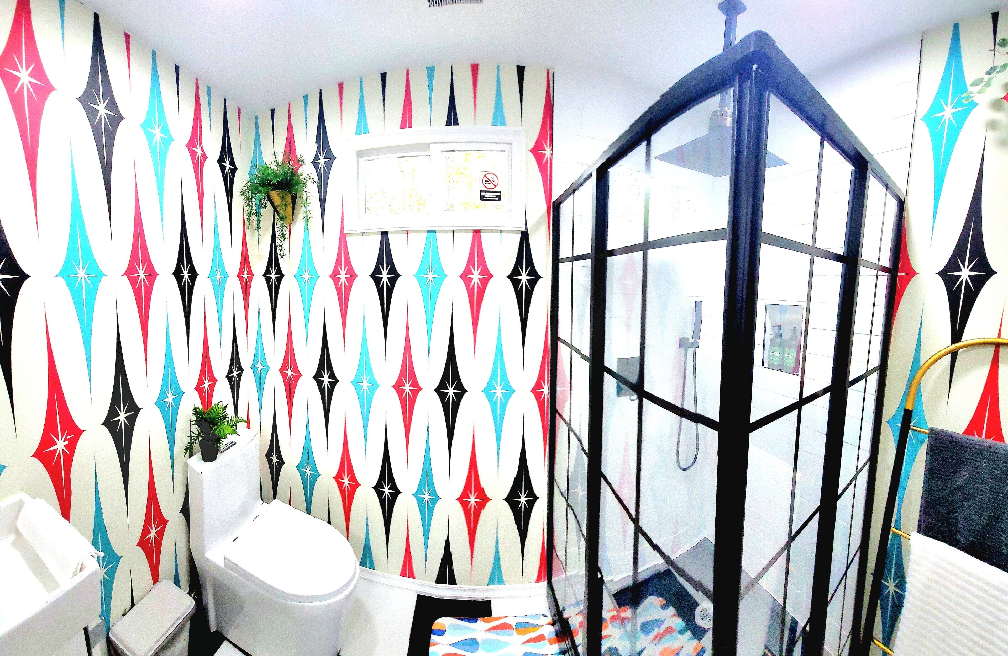A bathroom with a colorful geometric wall pattern, a white sink, a white toilet, and a glass-enclosed shower area.
