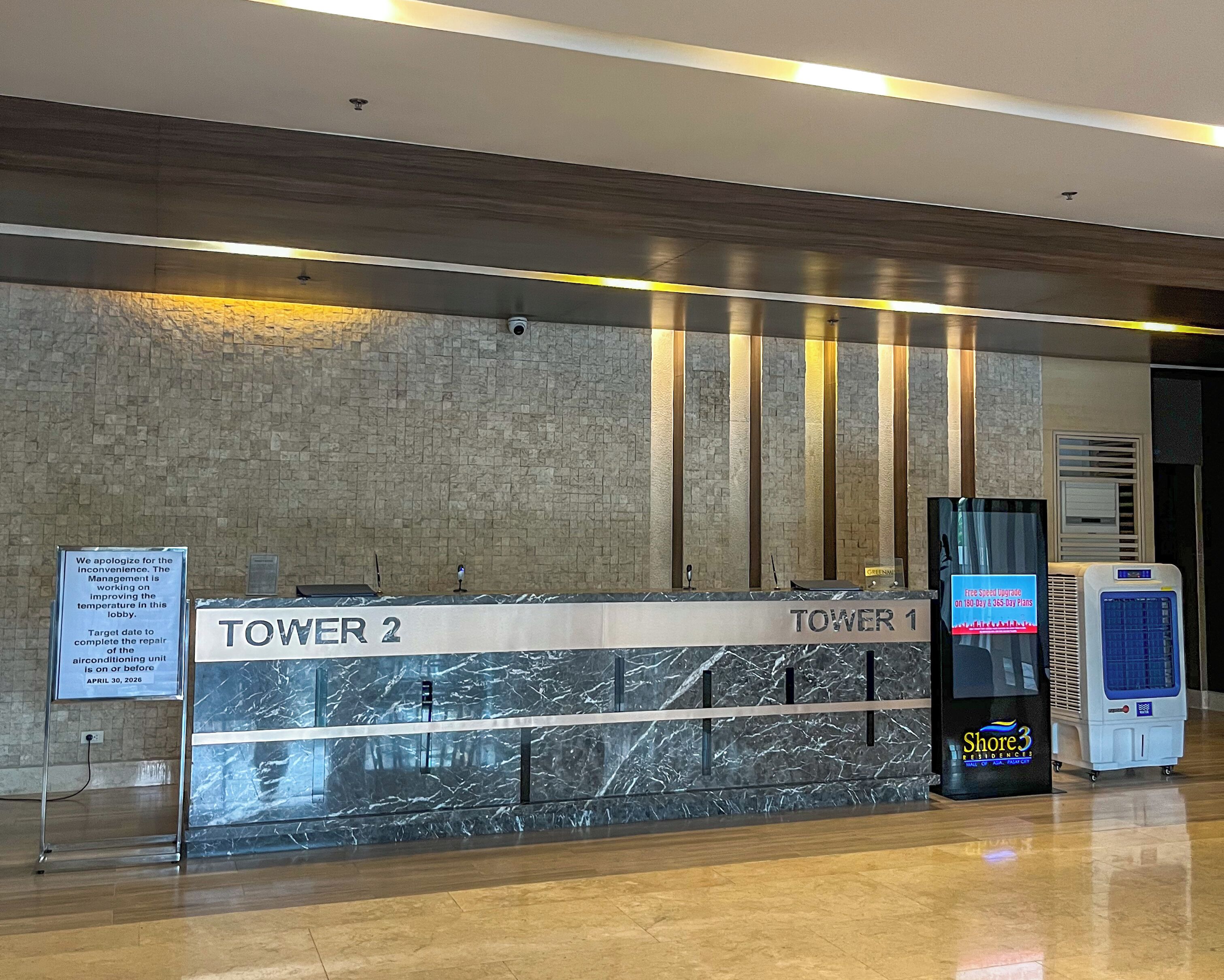 Ein Hotelrezeptionstresen mit Schildern für 'TOWER 1' und 'TOWER 2', einer Marmorplatte, einem digitalen Kiosk und Klimaanlagen.