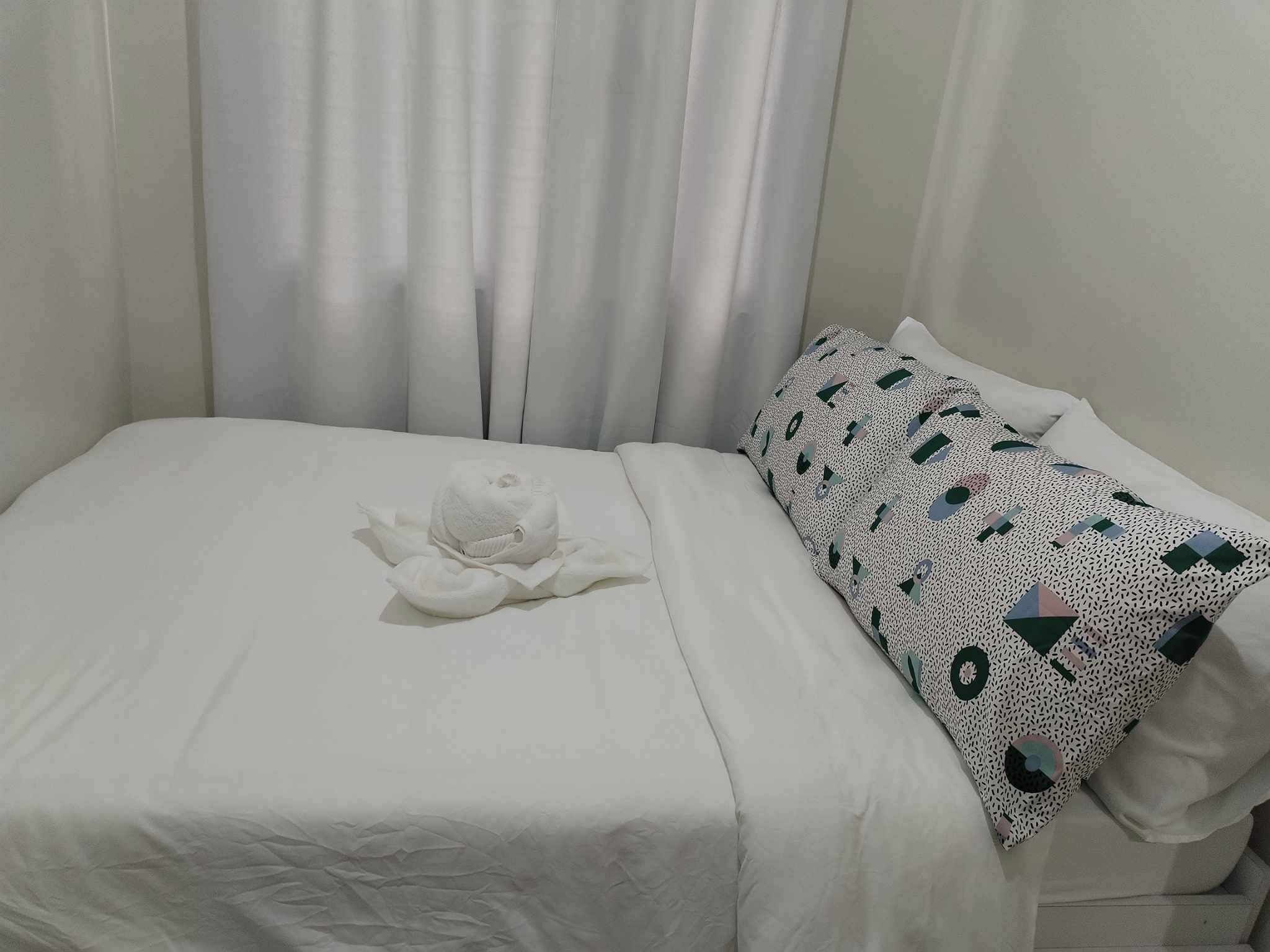 A neatly made bed with a towel and a patterned pillow in a room with white curtains.