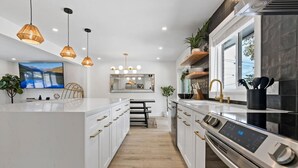 A modern kitchen with white cabinetry, a central island, pendant lighting, and a large window. - Arvada Olde Town Oasis Accomodates 24 People (Arvada)
