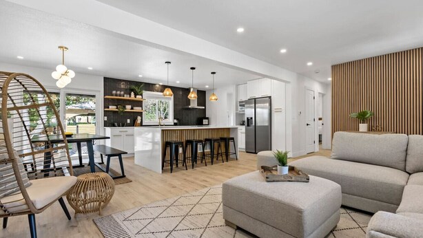 A modern kitchen with white cabinets, a central island, and pendant lighting. There is a dining area with a round table and chairs, and a living room with a sofa and ottoman. - Arvada Olde Town Oasis Accomodates 24 People (Arvada)
