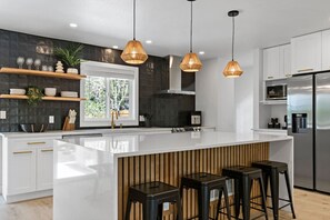 Une cuisine moderne dotée d’un grand îlot central, de luminaires suspendus et offrant une vue sur l’extérieur.
