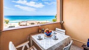 A balcony with a table set for two, overlooking a beach with clear blue water and a few buildings in the distance. - Bella Vista by Interhome (Algajola)