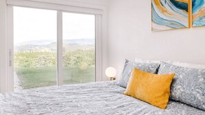Un dormitorio con un amplio ventanal corredizo que da a un balcón, una cama con ropa de cama estampada y una obra de arte colgada en la pared.