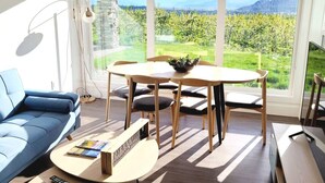 Una sala de estar moderna con un sofá azul, una mesa de comedor de madera con cuatro sillas y un gran ventanal que ofrece vistas a la vegetación y montañas.