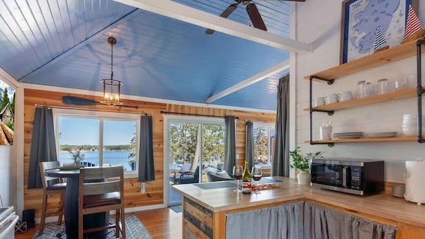 A spacious room with a wooden ceiling, a dining area with a table and chairs, a kitchen area with a microwave, and large windows offering a view of the outdoors.