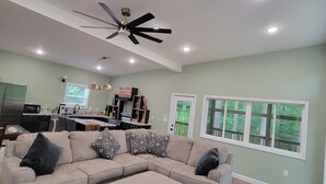 A modern living room with a sectional sofa, ottoman, ceiling fan, recessed lighting, and a view of the outdoors through large windows.