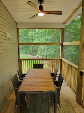 Une terrasse extérieure couverte, dotée d’une table et de chaises en bois, d’un ventilateur de plafond et offrant une vue sur une forêt.