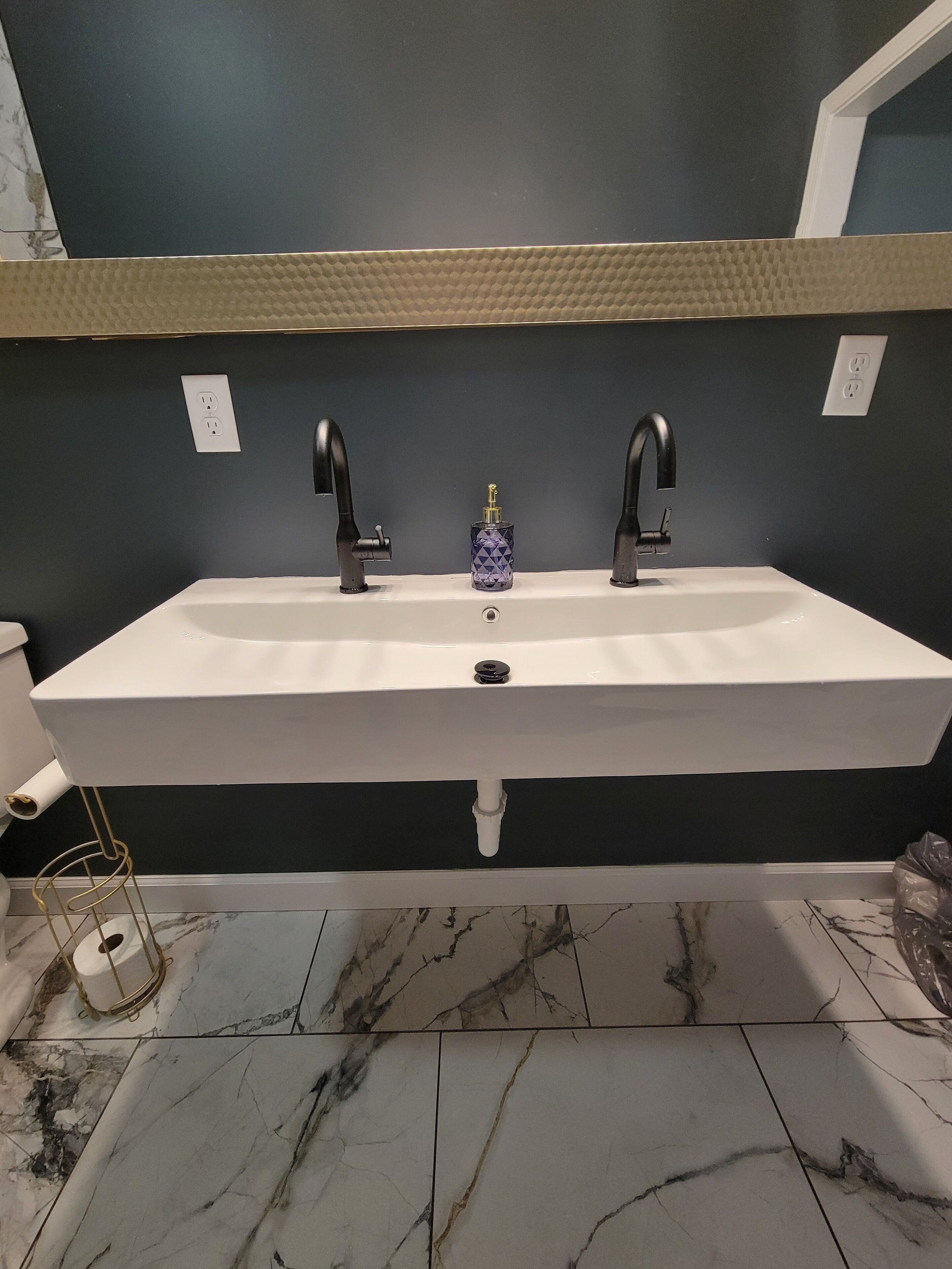 Un baño con un lavamanos blanco, dos grifos negros y suelo de mármol.