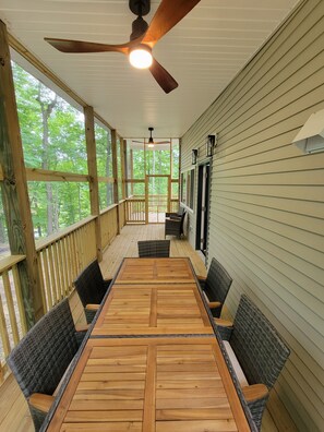 Une terrasse extérieure couverte, dotée d’une table et de chaises en bois, d’un ventilateur de plafond et de grandes fenêtres offrant une vue sur les arbres environnants.