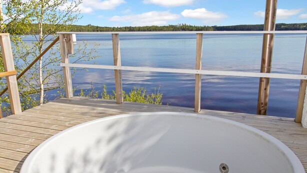 Un jacuzzi en una terraza de madera con vista a un lago tranquilo y a la orilla arbolada.