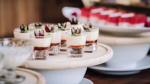 Una mesa de postres con copas de vidrio pequeñas que contienen postres en capas, adornadas con flores comestibles, y un plato de postre más grande al fondo.