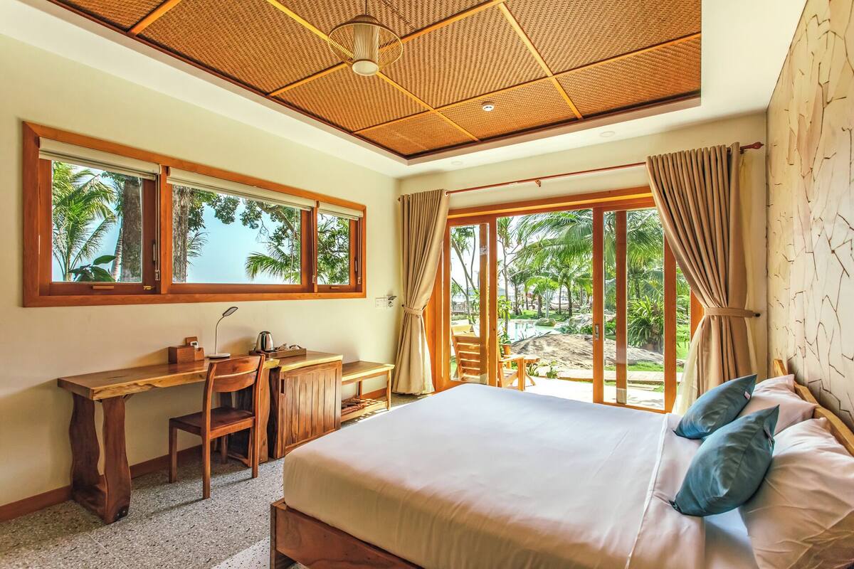 A bedroom with a large bed, a desk, and a view of palm trees through the windows.