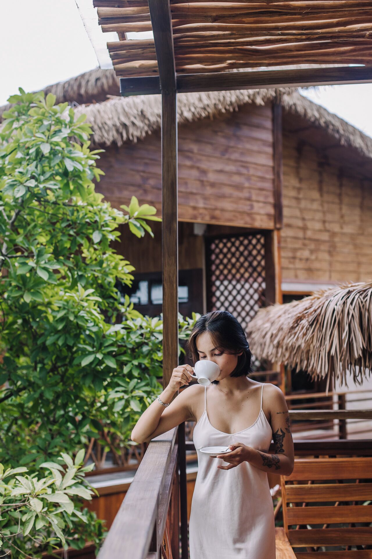 A woman in a white dress standing on a wooden deck with a thatched-roof building in the background.